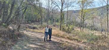 Un couple se tient sur un chemin de terre dans une forêt, entouré d'arbres et de nature, profitant d'une journée paisible et ensoleillée en plein air au milieu d'un terrain vallonné.