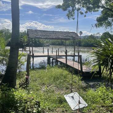 A tranquil riverside scene featuring a wooden dock with a thatched roof, lush greenery, a palm tree, a simple swing, and a calm, clear sky.