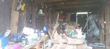 This image shows a cluttered wooden shed filled with various tools, equipment, containers, wood planks, bags, and boxes, creating a chaotic yet industrious workspace atmosphere.
