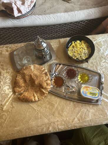 Ein traditioneller Frühstückstisch mit Teeservice, Gläsern, Fladenbrot, Eiern, verschiedenen Marmeladen, Olivenöl, Butter und Utensilien, gedeckt auf einer gemusterten Tischdecke. Gemütliches Ambiente.