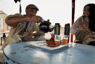 Ein Mann gießt fröhlich Tee aus einer Teekanne in Gläser auf einem Tablett, während eine Frau neben ihm lächelt. Die Umgebung ist zwanglos und einladend.