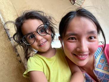 A imagem mostra uma jovem com óculos e camisa amarela, posando com uma mulher adulta em frente a uma parede bege, ambas sorrindo para a câmera.