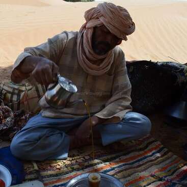La imagen muestra a un hombre sentado en una alfombra en el desierto, sirviendo té de una tetera metálica a un pequeño vaso de cristal.