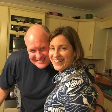 The image depicts a warm moment between a man and woman in a kitchen, with the woman\'s shoulder-length brown hair and blue-and-white top standing out against the beige cabinets and countertops.