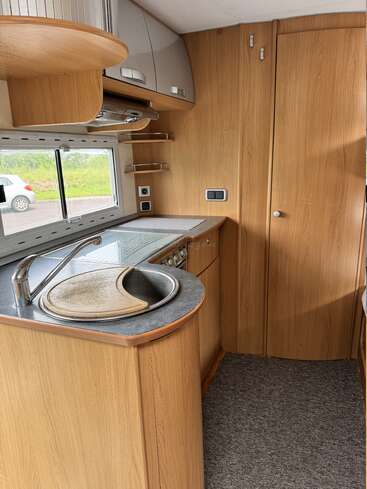 Das ist eine kompakte Wohnmobilküche mit hellen Holzschränken, grauen Arbeitsplatten, einem runden Spülbecken, einem Kochfeld, Hängeschränken, Regalen und einer Tür neben einem Fenster.