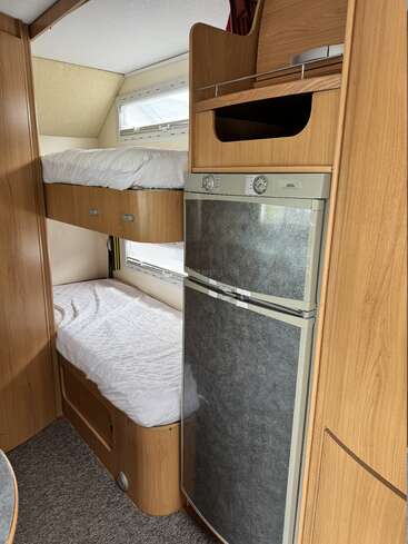 Dieses Bild zeigt einen gemütlichen Wohnmobil-Innenraum mit Etagenbetten aus Holz, einem kompakten Kühl-Gefrierkombination, Stauraum an der Decke, kleinen Fenstern und grauem Teppichboden, was einen aufgeräumten, zweckmäßigen Wohnraum schafft.