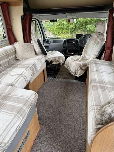 Dieses Bild zeigt das gemütliche Interieur eines Wohnmobils mit karierten Sitzbezügen, beigem Teppich, Kissen, Holzvertäfelung, Vorhängen und einem Blick auf die grüne Landschaft draußen.