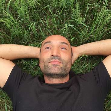 The image depicts a man lying on his back in a field of grass, with his arms raised above his head and wearing a black shirt.