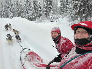 Two people bundled in red winter gear smile during a snowy dog sled ride through a pristine forest, surrounded by tall, snow-covered trees and happy huskies.