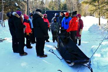A group of people dressed in winter clothing gathers around a sled in a snowy forest, possibly learning about dog sledding or preparing for an outdoor adventure.