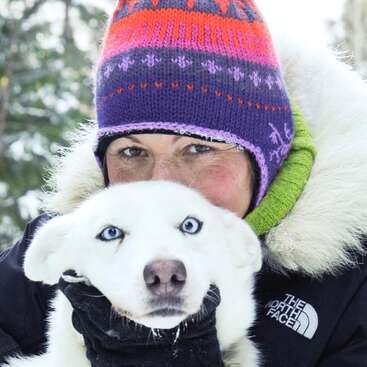 A person wearing a colorful winter hat and warm jacket hugs a white dog with striking blue eyes, outdoors in a snowy, forested setting, radiating warmth.