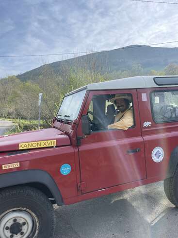 Das Bild zeigt einen Mann in einem roten Land Rover Defender mit weißem Dach, der am Straßenrand vor einer malerischen Kulisse aus Bäumen und Hügeln parkt.