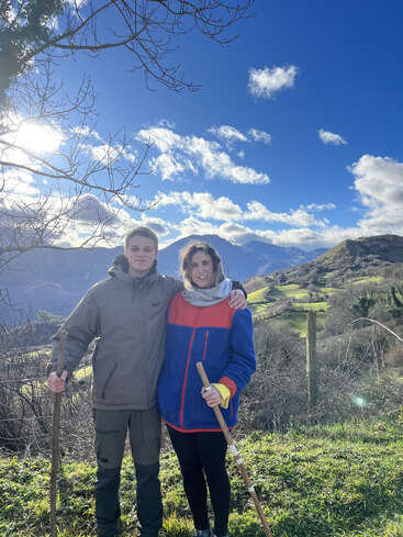 Das Bild zeigt einen jungen Mann und eine Frau, die mit Wanderstöcken in der Hand auf einer Wiese stehen, mit Bergen und einem blauen Himmel mit Wolken im Hintergrund.