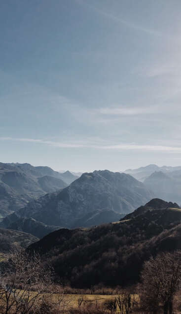 Das Bild zeigt eine ruhige Bergkette mit üppigem Grün und Bäumen vor einem blauen Himmel mit Wolkenfetzen, die ein Gefühl von natürlicher Schönheit und Ruhe hervorrufen.