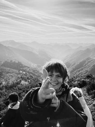 Das Bild ist eine Schwarz-Weiß-Fotografie, die eine Frau zeigt, die vor einer Berglandschaft den Daumen nach oben streckt. Die Frau ist der Hauptfokus.