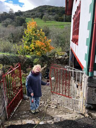 Das Bild zeigt eine Frau, die ein rotes Tor vor einem Haus malt, vor einer malerischen ländlichen Kulisse mit sanften Hügeln und Bäumen.