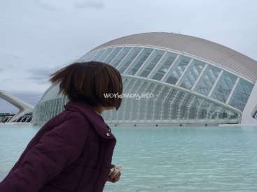 The image depicts a woman in a purple coat standing in front of a large glass building, with a body of water in the foreground.
