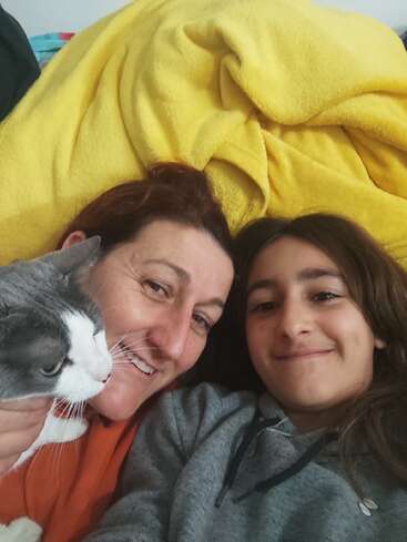 The image shows a woman and a girl lying on a yellow blanket, with a cat\'s head in the foreground, both smiling at the camera.