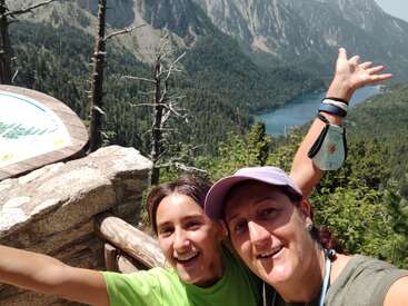The image shows two women posing for a selfie in front of a mountainous landscape with a lake and trees, with one woman\'s arm raised in the air.