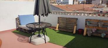 This rooftop terrace features artificial grass, a pallet lounge chair with a cushion, a round table with a closed umbrella, wooden planters, and cityscape views.