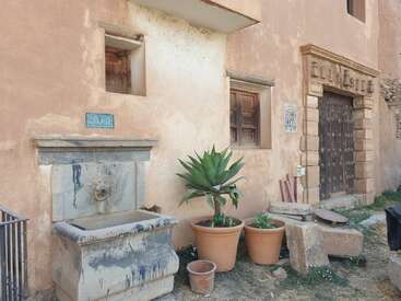 L'image représente un bâtiment usé par les intempéries, de couleur pêche, avec une fontaine en pierre, des plantes en pot et une porte marron foncé, ce qui lui confère un charme et un caractère rustiques.