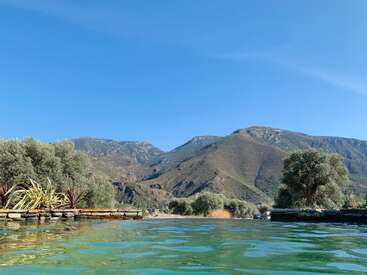 L'image représente un paysage serein avec une piscine, des arbres et des montagnes sous un ciel bleu clair, évoquant un sentiment de tranquillité et de beauté naturelle.