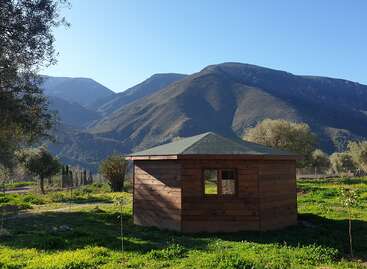 L'image représente une petite cabane en bois avec un toit vert, située dans un champ verdoyant entouré d'arbres et de montagnes à l'arrière-plan, par une journée ensoleillée.
