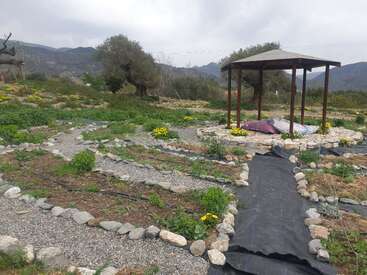 L'image représente un jardin serein avec un belvédère, avec un chemin sinueux, des rochers et des plantes, sur fond d'arbres et de montagnes sous un ciel couvert.