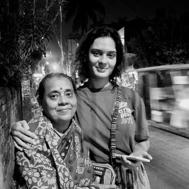 L'image représente un homme et une femme debout dans une rue la nuit, la main de la femme posée sur l'épaule de l'homme, tous deux regardant l'appareil photo.
