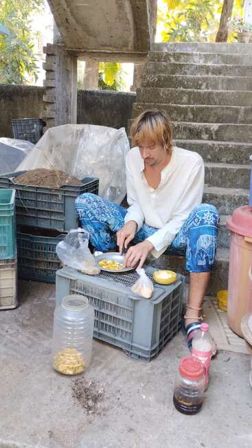 L'image représente un homme assis sur des marches en béton, préparant de la nourriture sur une caisse en plastique au milieu de divers conteneurs et caisses, avec un environnement extérieur serein à l'arrière-plan.