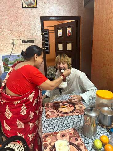 L'image représente une femme en sari rouge nourrissant un homme en chemise blanche à une table avec de la nourriture et des boissons, dans un cadre domestique.