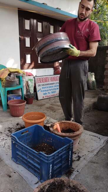 Un homme se tient dans une cour, tenant un grand pot en plastique, entouré de matériel de jardinage et d'un panneau de la Fondation Lieberearth.