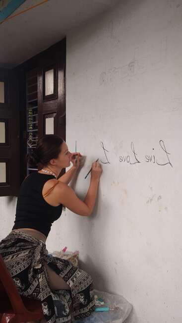 Une femme aux cheveux noirs, vêtue d'un débardeur noir et d'un pantalon à motifs, est assise sur une chaise rouge et écrit sur un mur blanc avec un stylo bleu.