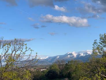 La imagen representa un paisaje sereno con una cordillera nevada, frondosos árboles y una pequeña ciudad enclavada en el valle, sobre un cielo azul con nubes.