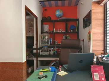 Ein kleiner Büroraum mit einem Schreibtisch, einem Laptop, einem Stuhl, einem Wasserspender, Regalen mit Gegenständen, einem Globus auf dem Kopf und Sonnenlicht durch die Jalousien.