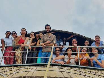 Um grupo diversificado de pessoas sorri e posa junto no convés de uma casa flutuante, desfrutando de uma experiência divertida ao ar livre sob um céu nublado, exalando alegria e camaradagem.