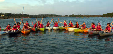 Um grupo de pessoas usando coletes salva-vidas vermelhos senta-se em caiaques coloridos em um lago, sorrindo e levantando os braços, desfrutando de um passeio aventureiro e divertido juntos.