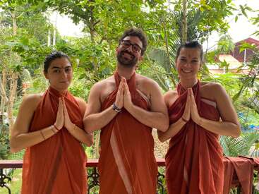 Três pessoas estão lado a lado ao ar livre, vestindo roupas laranja e fazendo um gesto respeitoso com as mãos juntas, sorrindo pacificamente em meio a árvores verdes exuberantes.