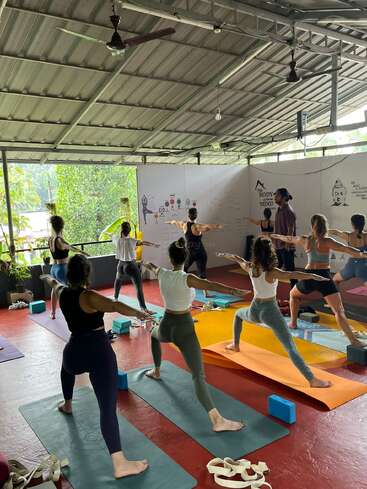 Um grupo de pessoas pratica ioga em um ambiente interno, em esteiras, orientadas por um instrutor. A luz natural, a vegetação externa, os pôsteres de instruções e os adereços de ioga criam uma atmosfera calma e concentrada.