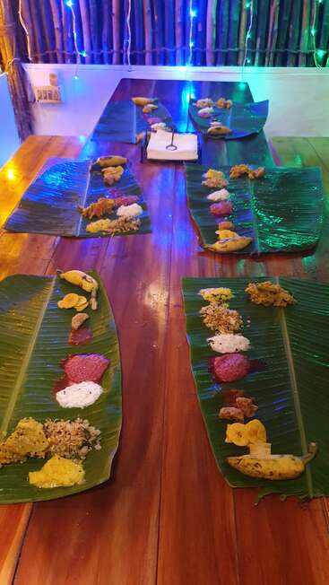 Folhas de bananeira servem como pratos, cuidadosamente dispostos em uma mesa de madeira, contendo pratos indianos tradicionais, chutneys, lanches e bananas sob luzes festivas e coloridas.