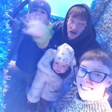 Uma família de quatro pessoas, toda agasalhada com roupas de inverno, sorri dentro de um espaço azul decorado com enfeites de Natal. As crianças estão usando chapéus; uma delas estende a mão na direção da câmera.
