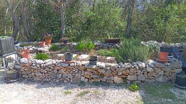 Esta imagem mostra um muro de pedra rústico de jardim com vários vasos de plantas e vegetação. Atrás do muro, há mais plantas, árvores e uma vegetação exuberante.