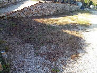 Uma área de cascalho com manchas de grama e folhas secas, delimitada por um muro de pedra. A luz do sol e as sombras cobrem o chão, criando um cenário sereno ao ar livre.