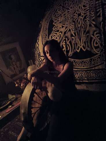 Une femme est assise dans une lumière tamisée et lunatique, tenant un grand tambour. Derrière elle, une tapisserie aux motifs celtiques complexes crée une atmosphère mystique et intime.