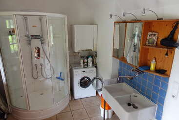 Esse banheiro contém uma cabine de chuveiro moderna, máquina de lavar roupa com porta aberta, pia quadrada, revestimento de azulejos azuis, prateleiras de madeira, espelho e vários produtos de higiene pessoal e eletrodomésticos.
