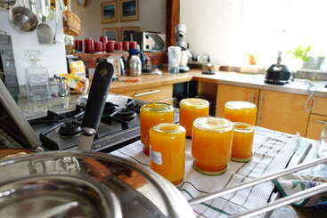 Esta cena brilhante de cozinha apresenta seis potes de geleia de laranja caseira esfriando de cabeça para baixo em uma toalha, cercados por vários utensílios de cozinha e eletrodomésticos sob a luz natural do sol.