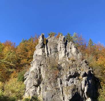 Um penhasco rochoso escarpado se ergue, cercado por árvores de outono vibrantes com folhas verdes, amarelas e laranjas. O céu acima é claro e intensamente azul.