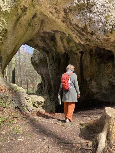 Uma pessoa com mochila vermelha e casaco cinza explora a entrada de uma caverna rochosa em uma floresta iluminada pelo sol, segurando um mapa ou livro, cercada pela natureza.