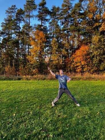 Uma pessoa joga badminton ao ar livre em um campo gramado, cercado por árvores de cores outonais e céu azul, desfrutando de um dia ativo ao ar livre e fresco.