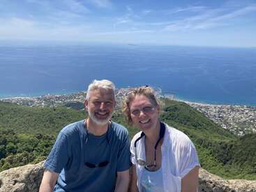 Um casal sorridente posa no topo de uma colina com uma cidade costeira deslumbrante e um oceano azul ao fundo, cercado por uma vegetação exuberante, sob um céu ensolarado.